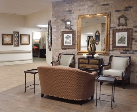 A cozy seating area in a senior living facility with a brown upholstered loveseat, two armchairs with light cushions, and two small side tables. Behind the seating is a brick wall adorned with framed artwork and a large gold-framed mirror. The floor is a combination of wood and carpet, and the hallway leading to other rooms is visible in the background.
