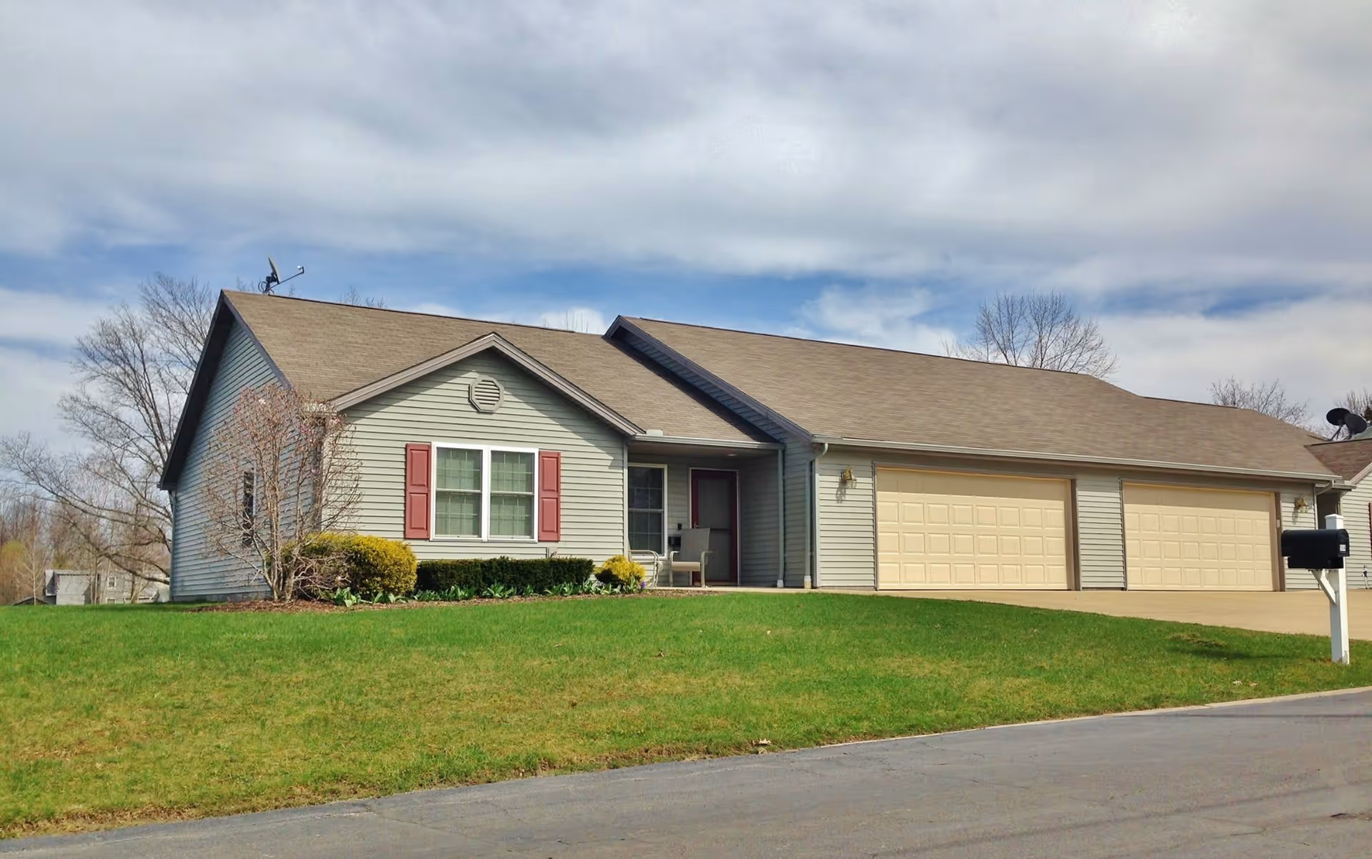 Single-story residential building with beige siding, a brown shingled roof, and three garage doors. The front yard has green grass and some bushes near the house. The sky is partly cloudy.