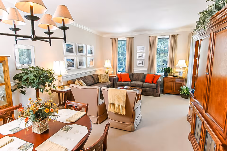A bright and cozy living room with two large windows covered by beige curtains, two brown sofas with orange and patterned cushions, two beige armchairs with a yellow throw blanket, a wooden coffee table, and several lamps providing warm lighting. A wooden dining table with floral centerpiece and placemats is visible in the foreground. The room is decorated with framed pictures on the walls and several green plants.