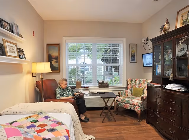 A cozy room with a bed covered by a colorful patchwork quilt and a cream knitted blanket. An elderly woman is sitting in a brown armchair reading a book. Next to her is a floral upholstered armchair and a small wooden folding table. A large window with white blinds lets in natural light. The room has wooden flooring, a lamp on a side table, framed pictures on the wall, and a dark wooden cabinet displaying decorative items and papers.
