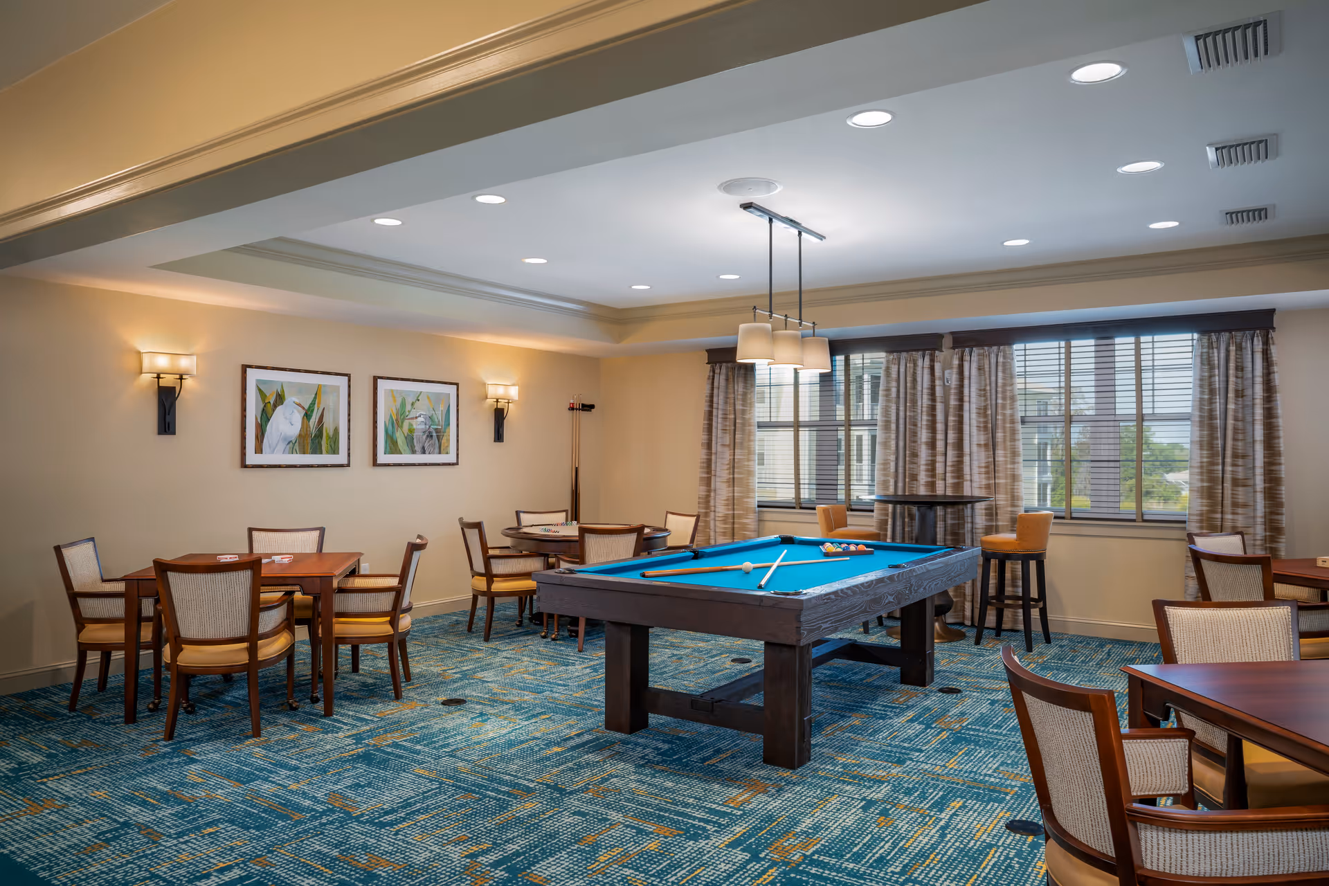 Bright interior recreation room with a pool table at center, multiple card tables and chairs, framed artwork on the wall, and large windows with curtains.