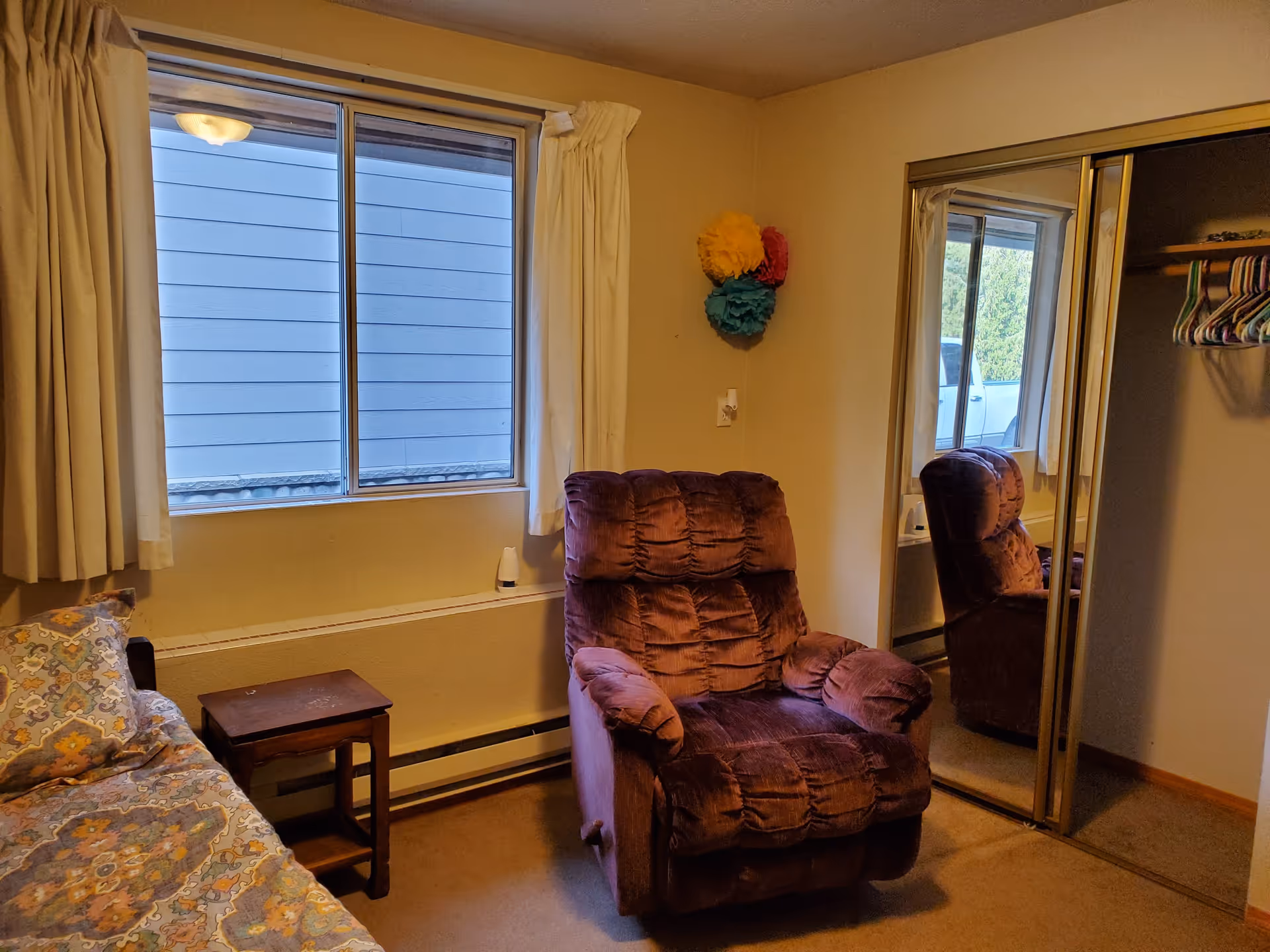 A cozy bedroom with a patterned bedspread on a bed, a small wooden side table, a plush purple recliner chair, a window with cream-colored curtains, and a closet with mirrored sliding doors reflecting the chair. Colorful paper decorations are mounted on the wall above the chair.