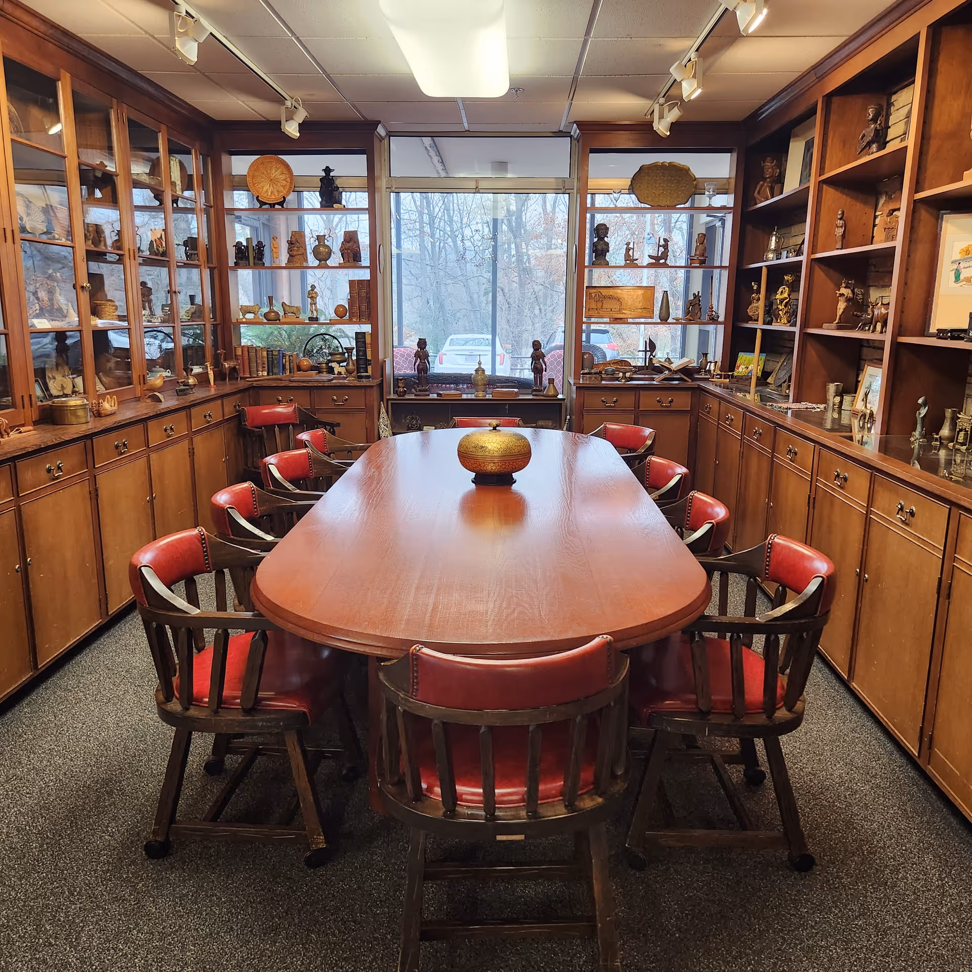A conference or meeting room with a long wooden table surrounded by ten wooden chairs with red cushions. The room has wooden cabinets and shelves filled with various decorative items, artifacts, and books. Large windows at the far end show a view of trees and parked cars outside.