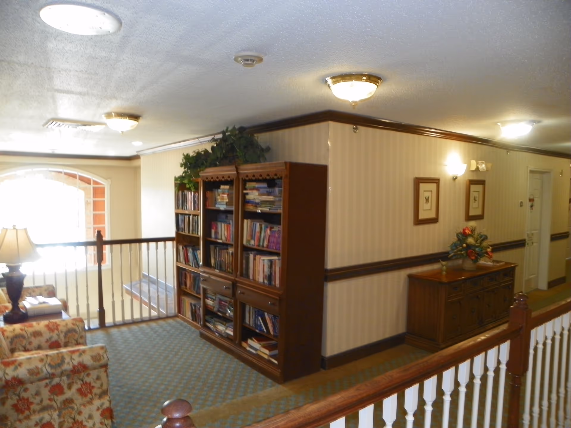 Interior hallway area of a senior living facility with a wooden bookshelf filled with books, a floral patterned armchair, a wooden sideboard with a flower arrangement, framed pictures on the wall, and ceiling lights. The area has carpeted flooring and a railing overlooking a lower level.