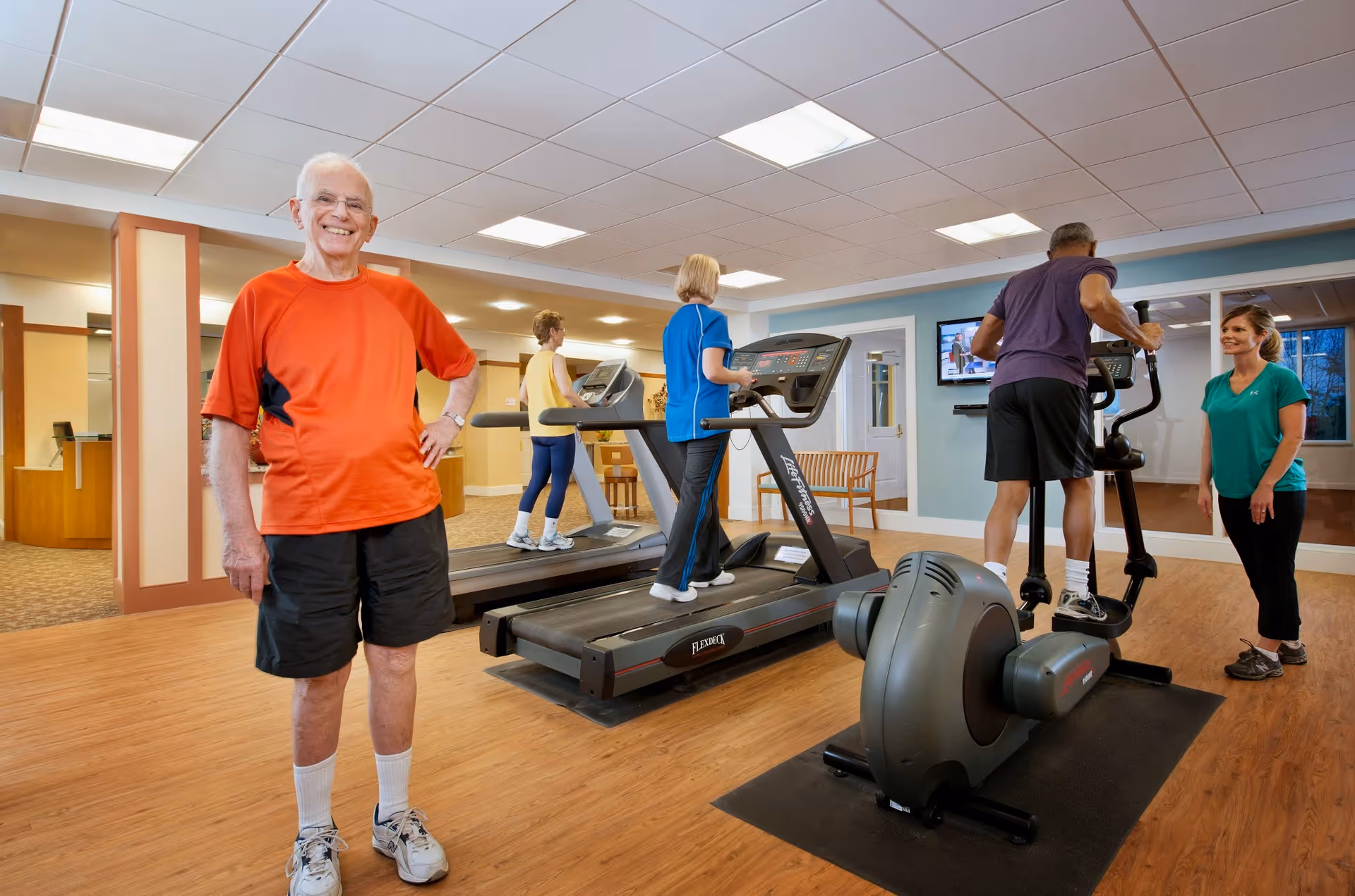 An indoor fitness room at North Hill Retirement Community with four seniors exercising on treadmills and an elliptical machine. A smiling elderly man in an orange shirt and black shorts stands in the foreground. A female instructor in a green shirt is assisting a man on the elliptical. The room has wood flooring, large mirrors, and bright ceiling lights.