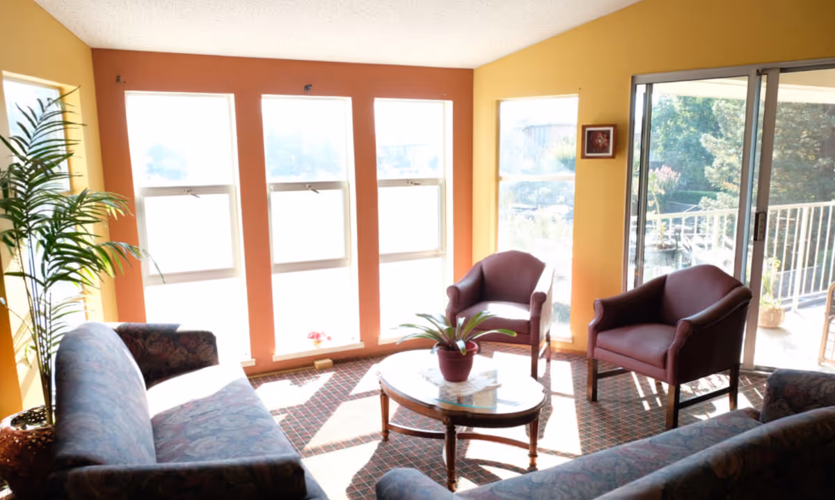 A bright living room with large windows allowing sunlight to fill the space. The room has two patterned sofas, two maroon armchairs, a wooden coffee table with a potted plant on it, and a sliding glass door leading to an outdoor balcony. The walls are painted yellow and orange, and there is a tall green plant in a pot near the window.