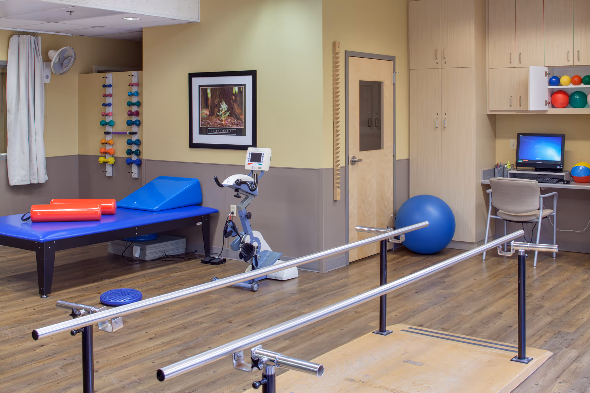 A physical therapy room with parallel bars for walking exercises, a stationary exercise bike, a blue therapy table with red foam rollers, a rack of colorful dumbbells, a large blue exercise ball, and a desk with a computer and chair. The room has wooden flooring, beige and gray walls, and a framed motivational picture on the wall.