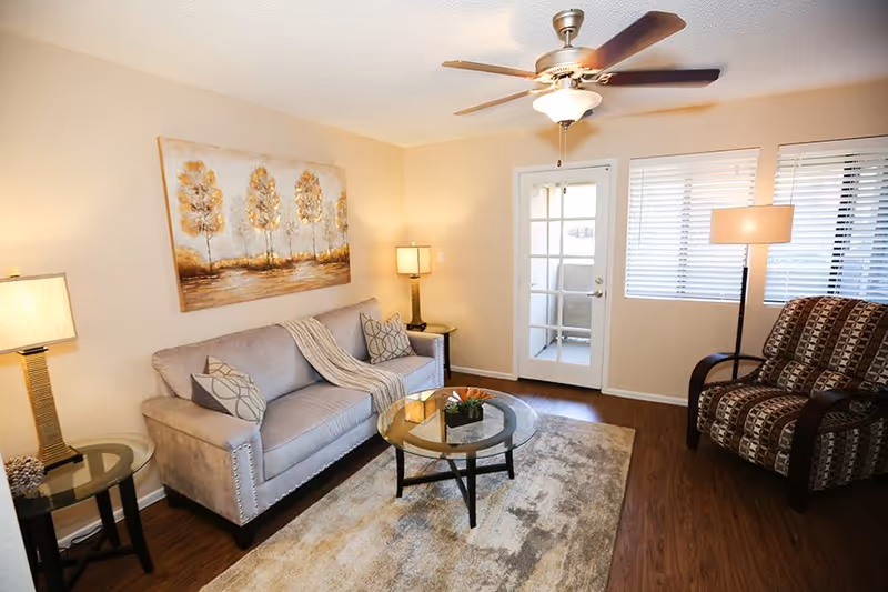 A cozy living room with a beige sofa adorned with patterned pillows and a throw blanket, a glass coffee table with a small plant centerpiece, a patterned armchair, two side tables with lamps, a ceiling fan with light, and a large painting of trees on the wall. There is a door with glass panels and windows with blinds letting in natural light.