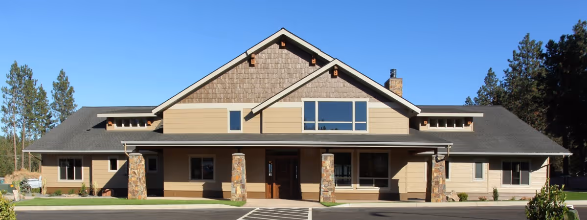 Front exterior view of a single-story building with a peaked roof, stone pillars supporting a covered entrance, beige siding, and large windows, surrounded by trees and a clear blue sky.