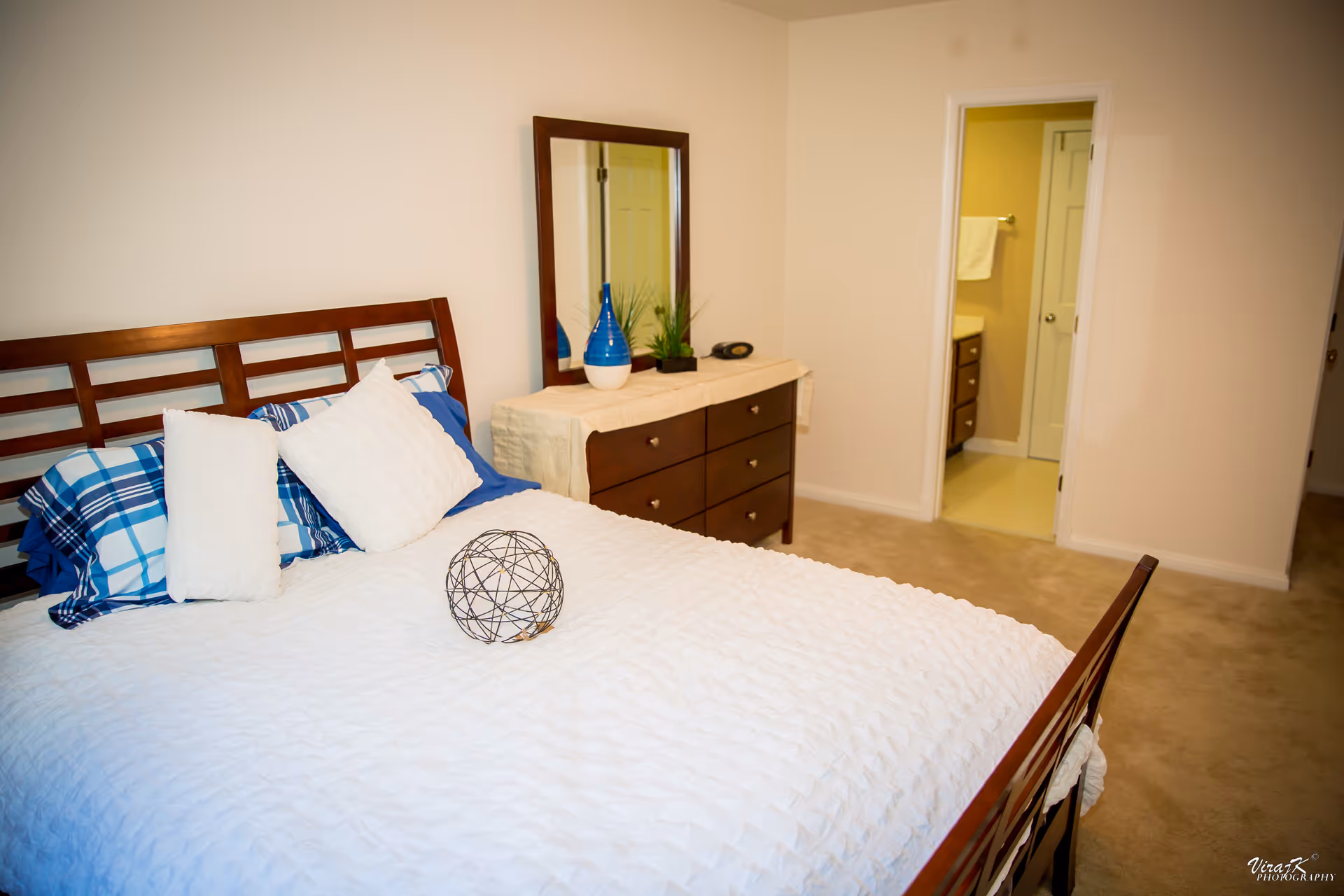 Neatly made bedroom with a wooden bed, pillows, dresser with mirror, and an open doorway to a bathroom.