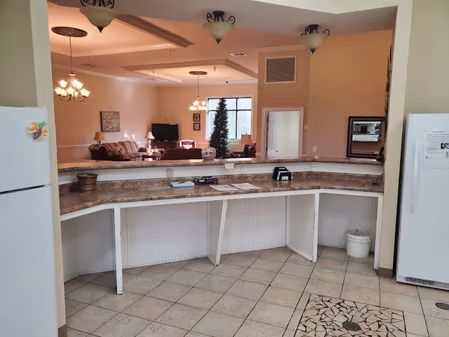 Interior view of a senior care facility showing a curved countertop with a granite-like surface and white cabinets underneath. In the background, there is a living room area with a sofa, a TV, a Christmas tree, and hanging light fixtures. Two white refrigerators are visible on either side of the countertop area.
