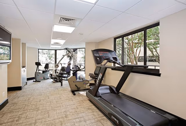 A fitness room with exercise equipment including a treadmill, stationary bike, and multi-function weight machine. The room has large windows letting in natural light and a wall-mounted TV.