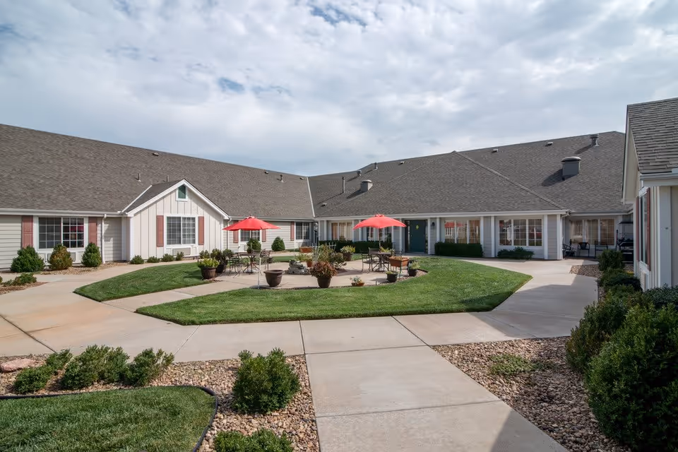 Outdoor courtyard area of a senior living facility with paved walkways, green grass, potted plants, and tables with red umbrellas. The building surrounding the courtyard has white siding, multiple windows, and a gray roof under a partly cloudy sky.
