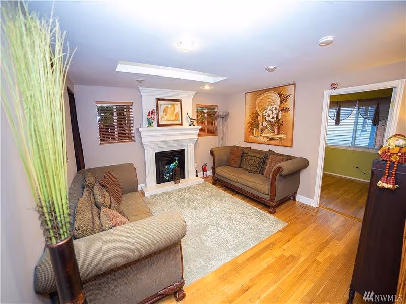 A cozy living room with two brown upholstered sofas facing each other, a white fireplace with decorative items on the mantel, a light-colored area rug on a wooden floor, and a large framed artwork on the wall. There are two small windows with wooden blinds and a tall vase with green plants in the foreground. An adjacent room with a window and green walls is visible through an open doorway.
