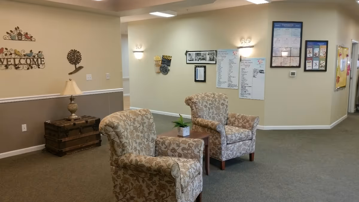 A cozy seating area in a senior living facility with two floral-patterned armchairs facing each other and a small wooden table with a potted plant between them. The walls are decorated with framed pictures, posters, and a welcome sign with colorful birds. A vintage-style trunk with a lamp on top is placed against the wall. The space is well-lit with wall sconces and overhead lights.