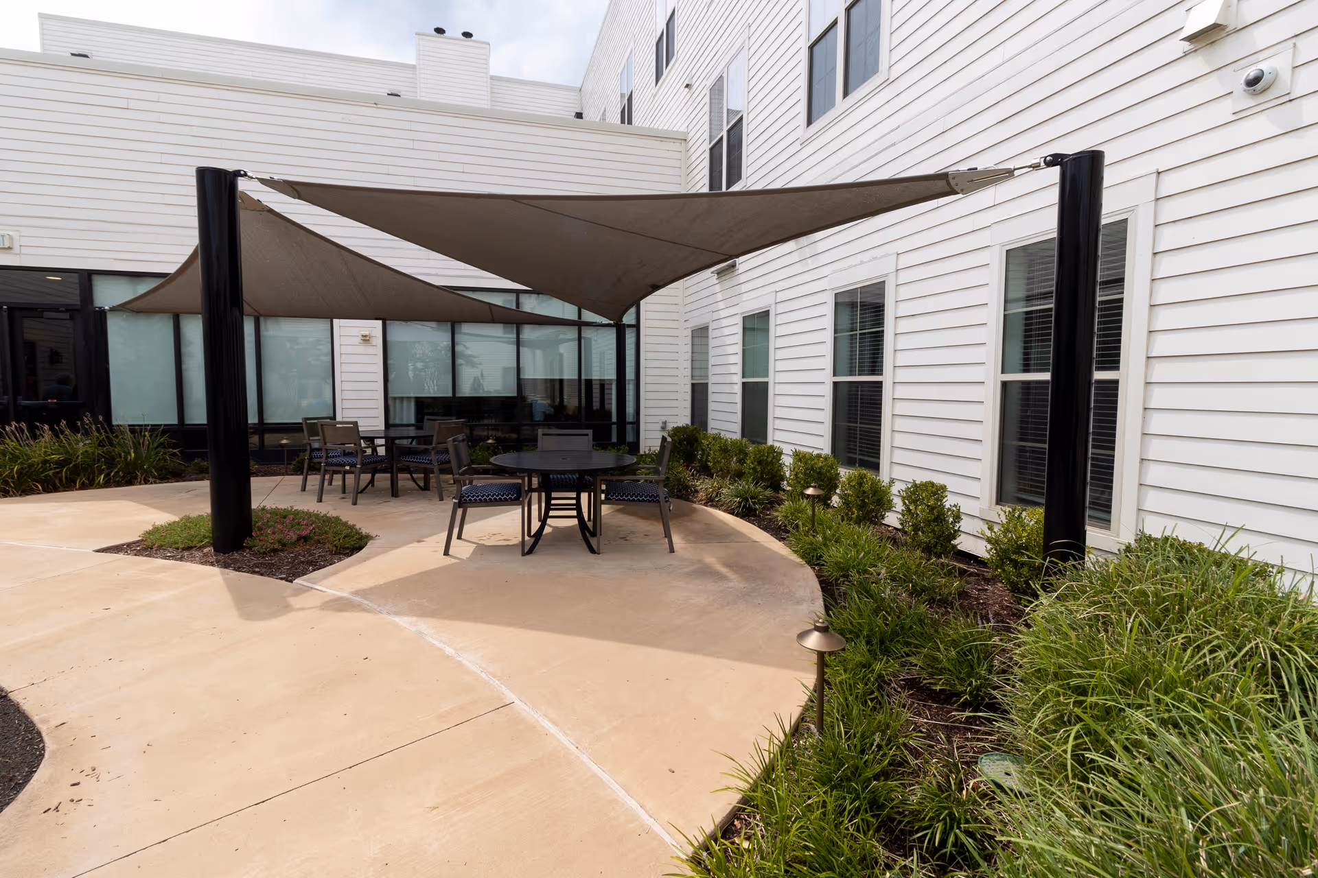 Outdoor patio area at StoryPoint Germantown with a concrete floor, several chairs and tables under a large triangular shade sail, surrounded by landscaped greenery and the white exterior walls of the building with multiple windows.