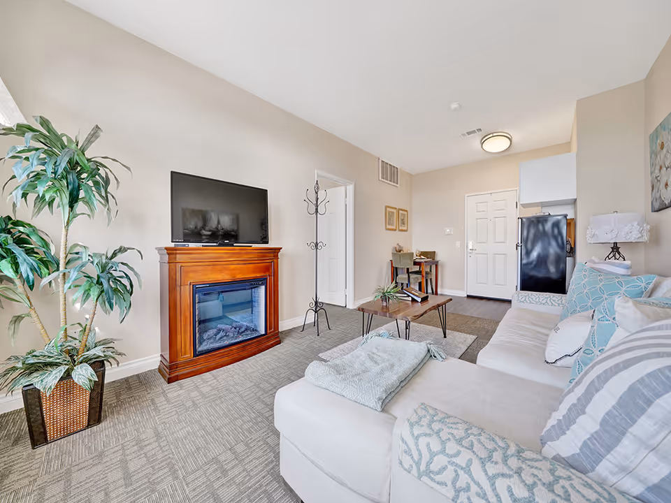 Bright furnished living room with a sectional sofa, TV on a wooden fireplace mantel, coffee table, and potted plant.