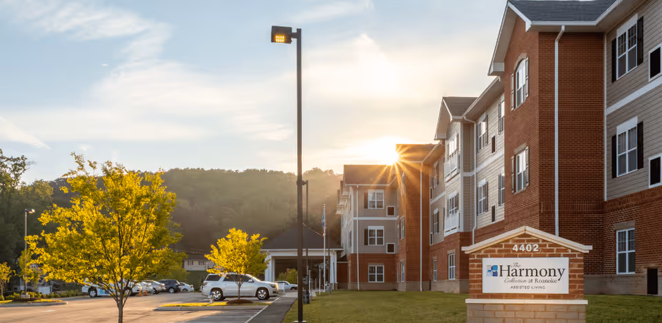 Exterior view of The Harmony Collection at Roanoke - Assisted Living facility during sunset, showing a multi-story brick and siding building, a parking lot with cars, trees with yellow leaves, and a sign with the facility name and address 4402.