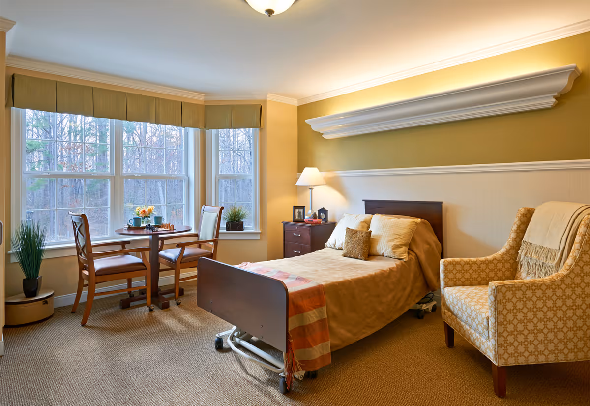 A cozy bedroom in a retirement facility with a single bed covered in beige bedding, a bedside table with a lamp and framed photos, a patterned armchair with a throw blanket, and a small round table with two chairs near large windows overlooking trees.