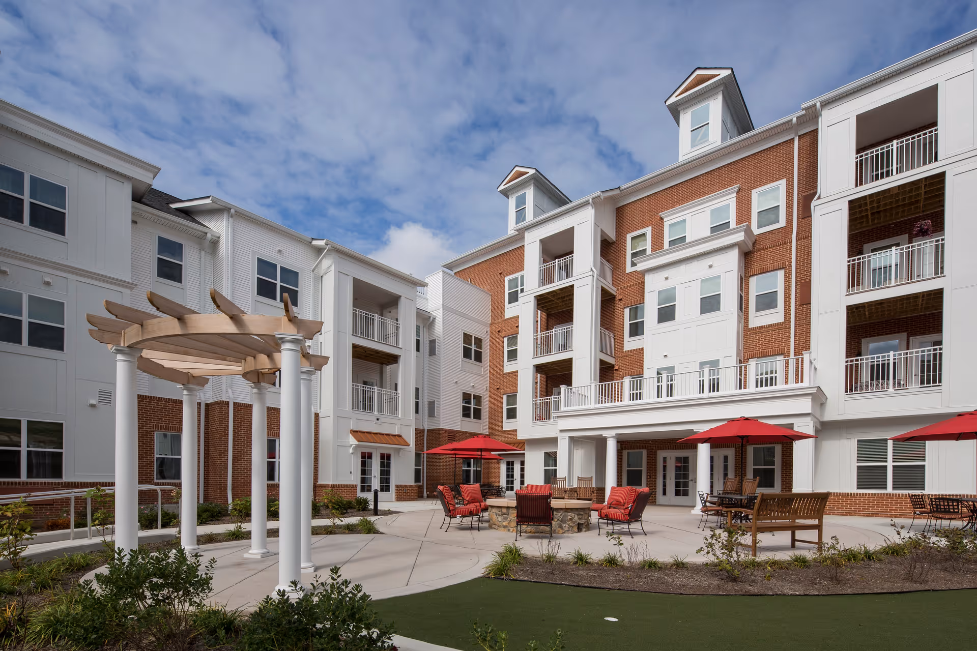 Outdoor courtyard area of a senior living facility with a multi-story building in the background. The courtyard features a pergola, several seating areas with red umbrellas, chairs, and benches arranged around a fire pit. The building has white and red brick exterior walls with balconies and large windows under a partly cloudy sky.