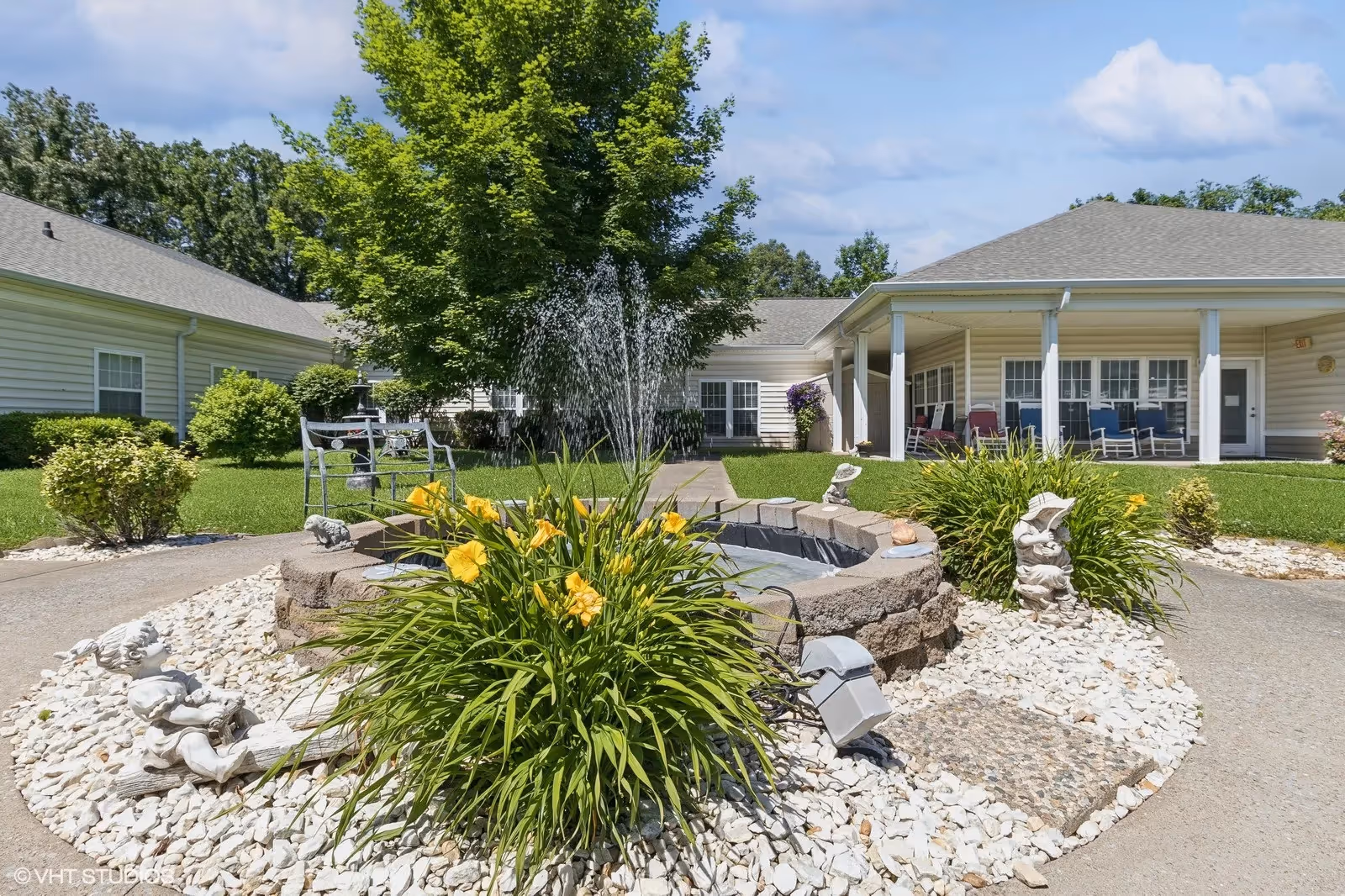 Outdoor courtyard area at The Bungalows at Mayfield featuring a small circular water fountain surrounded by yellow flowers and decorative garden statues. The courtyard is bordered by a paved walkway and green grass, with a large tree and single-story buildings with covered porches and rocking chairs in the background under a partly cloudy sky.