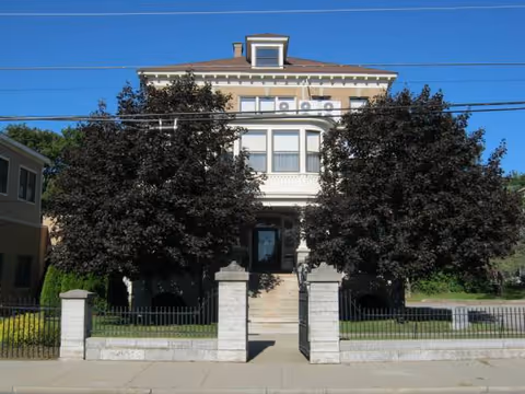 Front view of a two-story senior living home framed by large trees and a gated entrance.