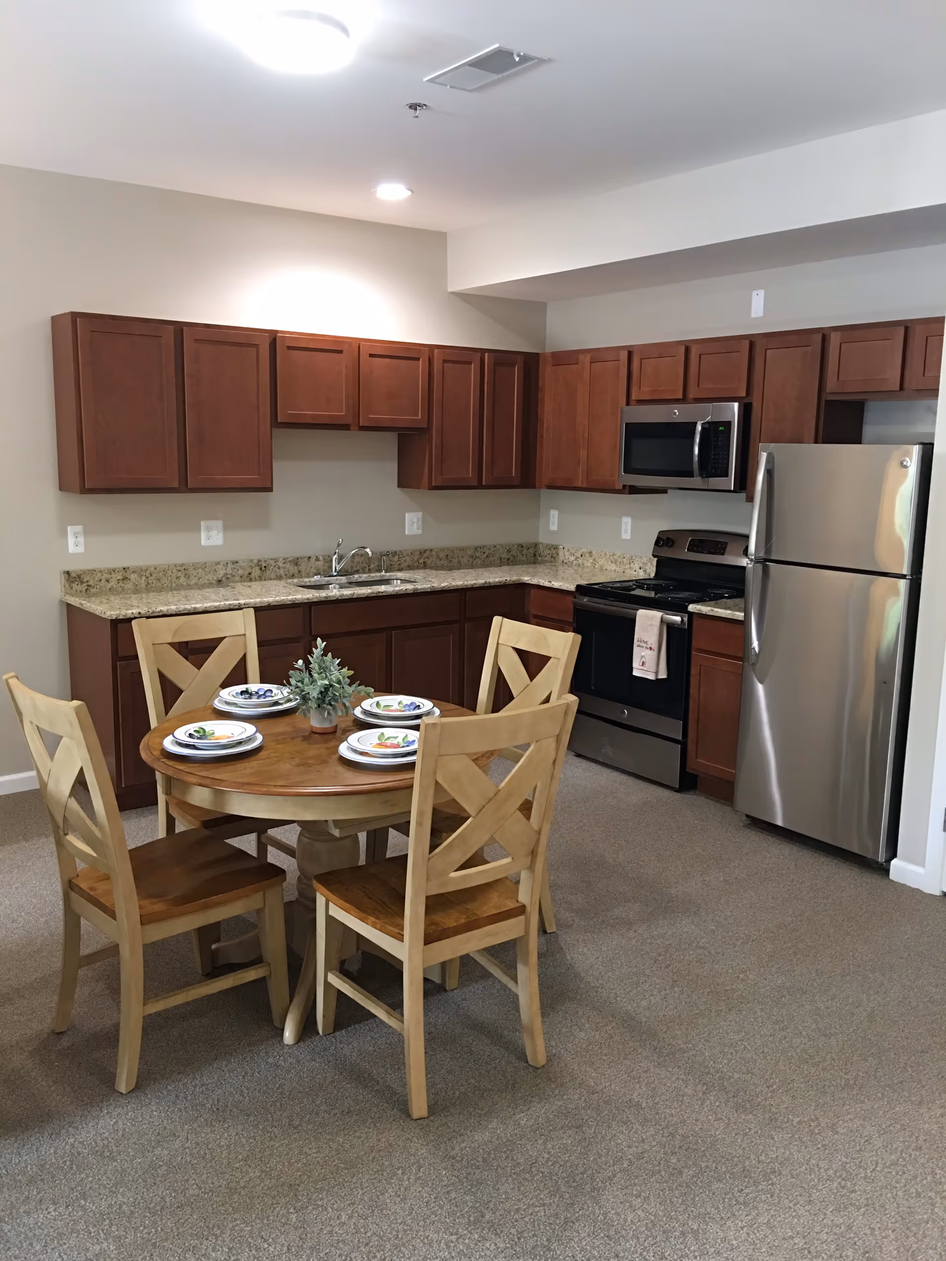 A kitchen area in a senior apartment featuring wooden cabinets with a granite countertop, a stainless steel refrigerator, stove, and microwave. In front of the kitchen is a round wooden dining table set with four chairs and four place settings, along with a small potted plant centerpiece.