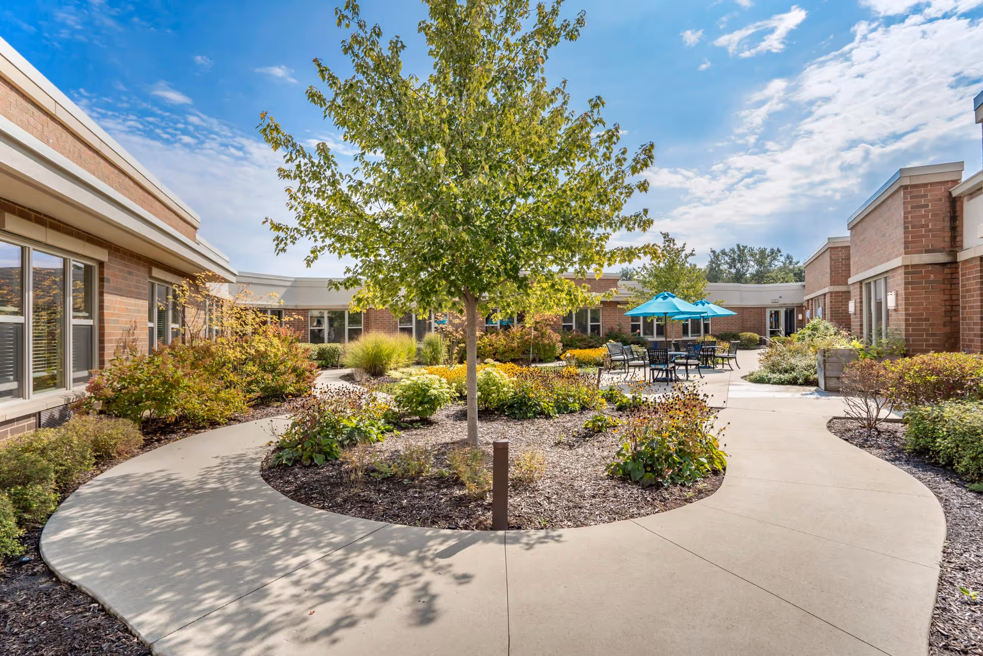 Outdoor courtyard area at The Sheridan at Green Oaks featuring a curved concrete walkway surrounding a landscaped garden with a tree and various plants. There are patio tables with blue umbrellas and chairs for seating, bordered by brick buildings under a partly cloudy sky.