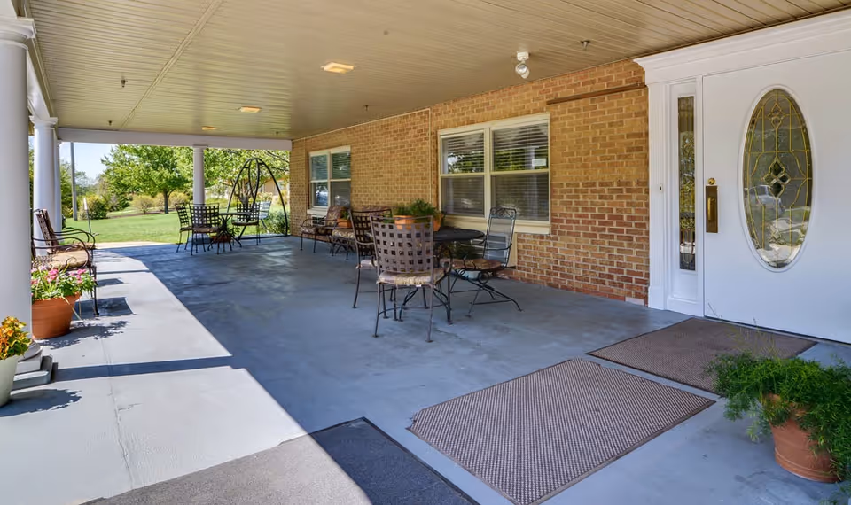 Covered front porch with outdoor tables and chairs, potted plants, and an oval-glass entry door on a brick building.
