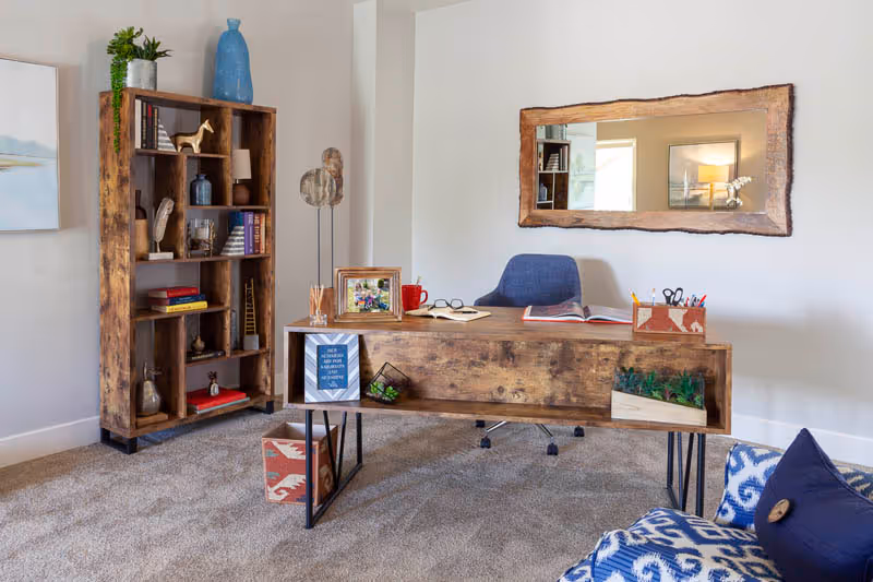 A cozy office space with a wooden desk and a blue office chair. The desk has an open book, a red mug, a photo frame, and a container with pens and scissors. Behind the desk is a large wooden-framed mirror on the wall. To the left, there is a wooden bookshelf with various decorative items and books. The room has beige carpet and light-colored walls, with a blue patterned armchair partially visible in the foreground.