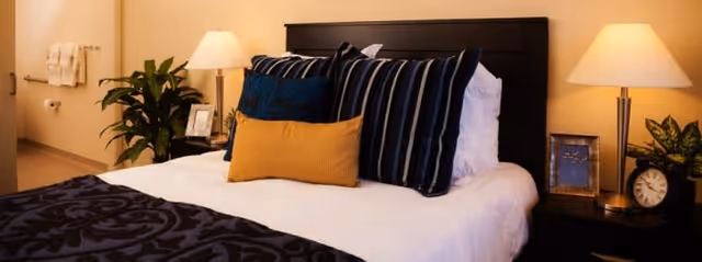 A neatly made bed with multiple pillows including striped and solid colored ones, flanked by two bedside tables each with a lamp. There is a plant on the left side near the wall and a clock and framed picture on the right bedside table. The room has warm lighting and a cozy atmosphere.