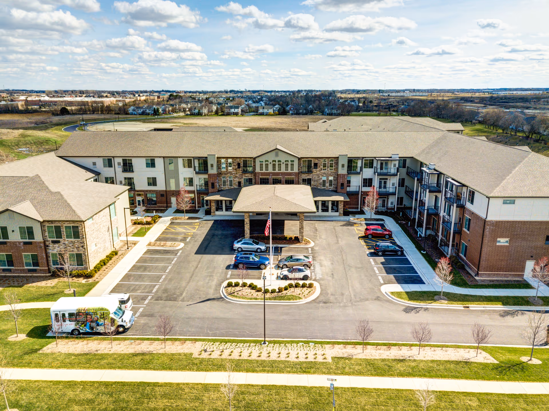 Aerial view of The Oaks at Algonquin Senior Living facility showing a large three-story building with a central entrance covered by a canopy. The building has a mix of brick and light-colored siding with balconies on the right side. There is a parking lot in front with several cars parked and a van on the left side. The surrounding area includes grassy lawns and a distant view of houses and open land under a partly cloudy sky.