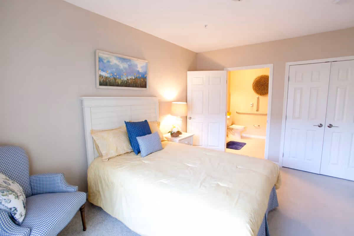 A bedroom with a white wooden headboard bed covered with beige bedding and blue pillows. To the left is a blue and white checkered armchair with a floral cushion. A nightstand with a lamp and a small plant is beside the bed. The room has beige walls and carpeted floor. An open door reveals a bathroom with a toilet, grab bars, and a blue bath mat. There is also a closed double-door closet in the room.