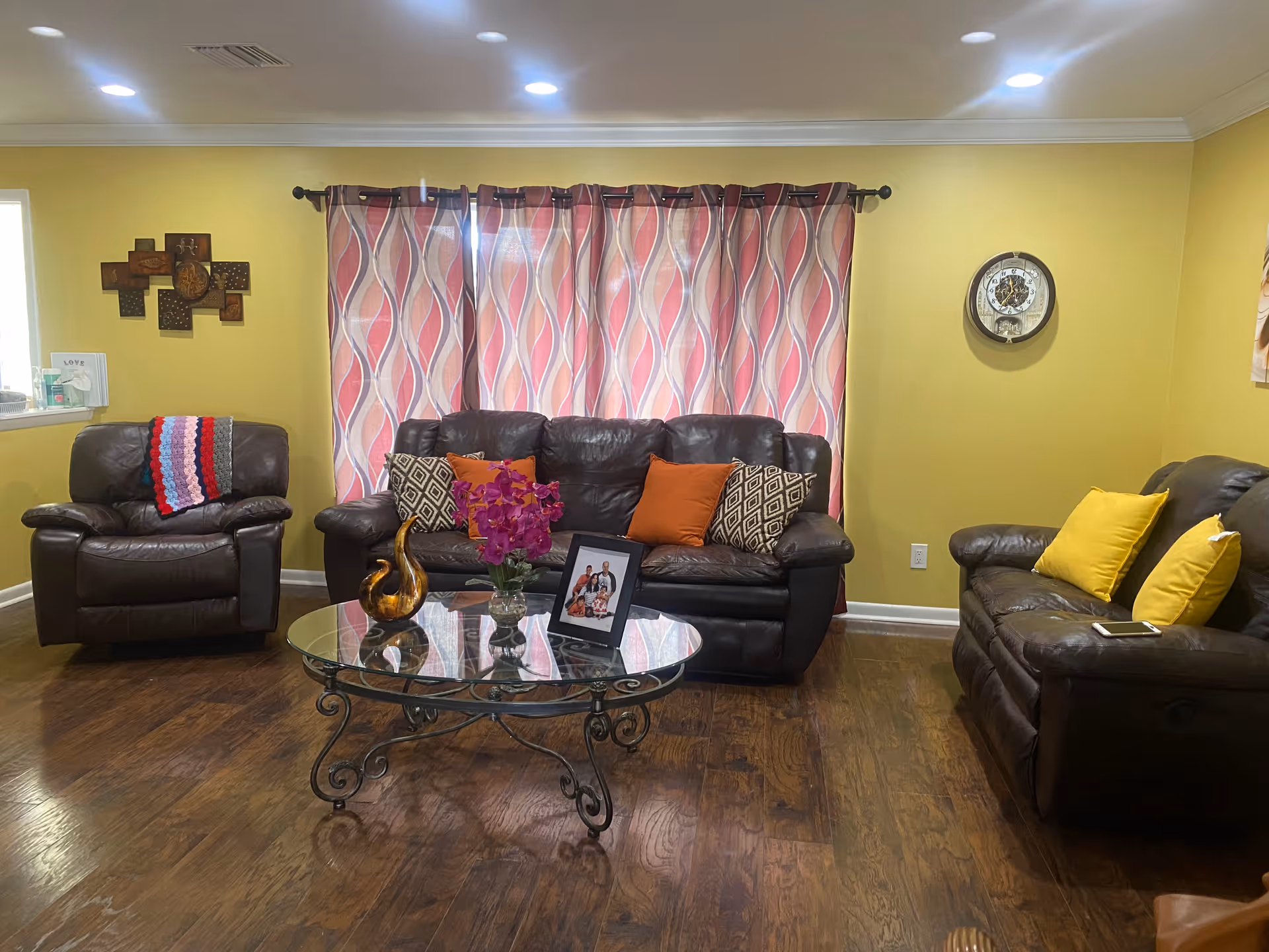 Cozy living room with dark leather sofas and armchairs, a glass coffee table with flowers and a photo, patterned curtains and yellow walls.