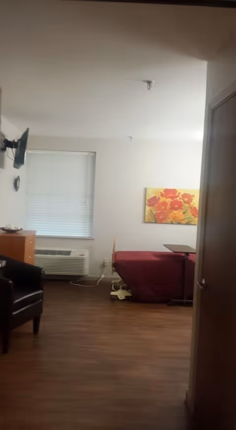 Small nursing facility room with a raised bed covered in a red blanket, an overbed table, wall-mounted TV, chair, and a floral painting on the wall.