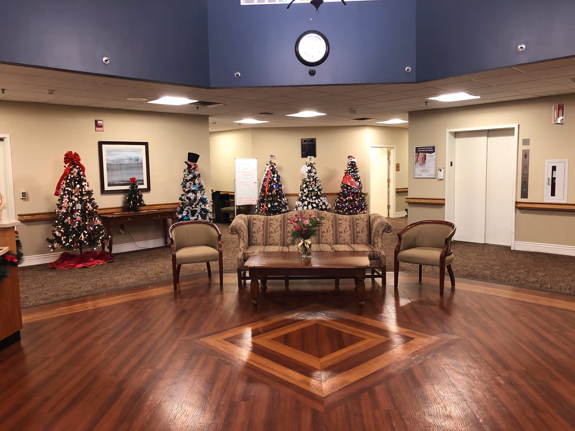 A spacious lobby area with polished wooden floors and a patterned wooden inlay in the center. There is a floral-patterned sofa and two armchairs arranged around a wooden coffee table with a vase of flowers. Behind the seating area, there are five decorated Christmas trees with various ornaments and ribbons. The walls are beige with a framed picture and a clock mounted on a blue upper wall section. An elevator and hallway doors are visible in the background.