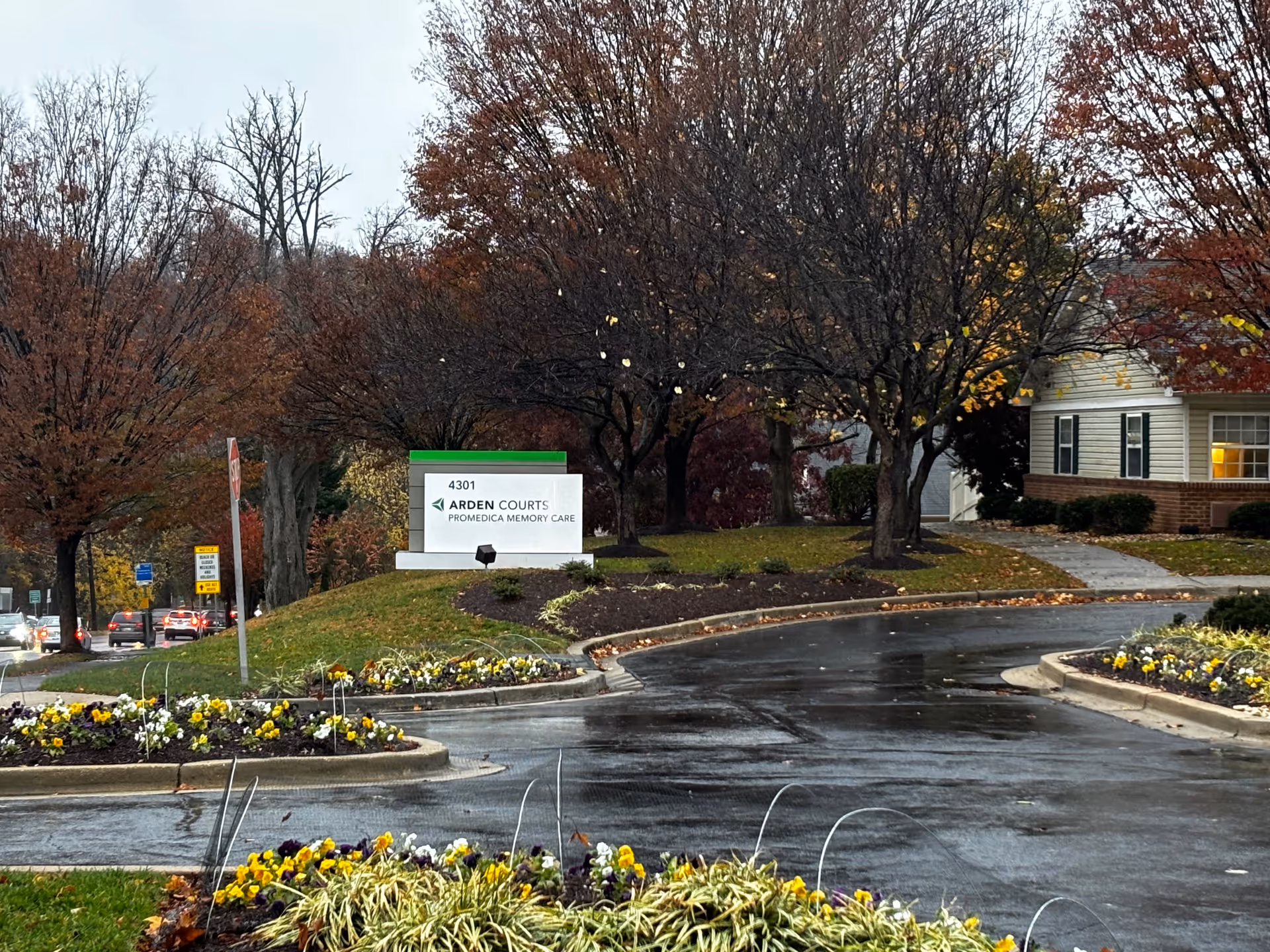 Entrance driveway to Arden Courts - ProMedica Memory Care Community with a sign displaying the facility name and address 4301. The scene includes landscaped flower beds with yellow and white flowers, a wet road, and trees with autumn foliage. A building with lit windows is partially visible on the right side.