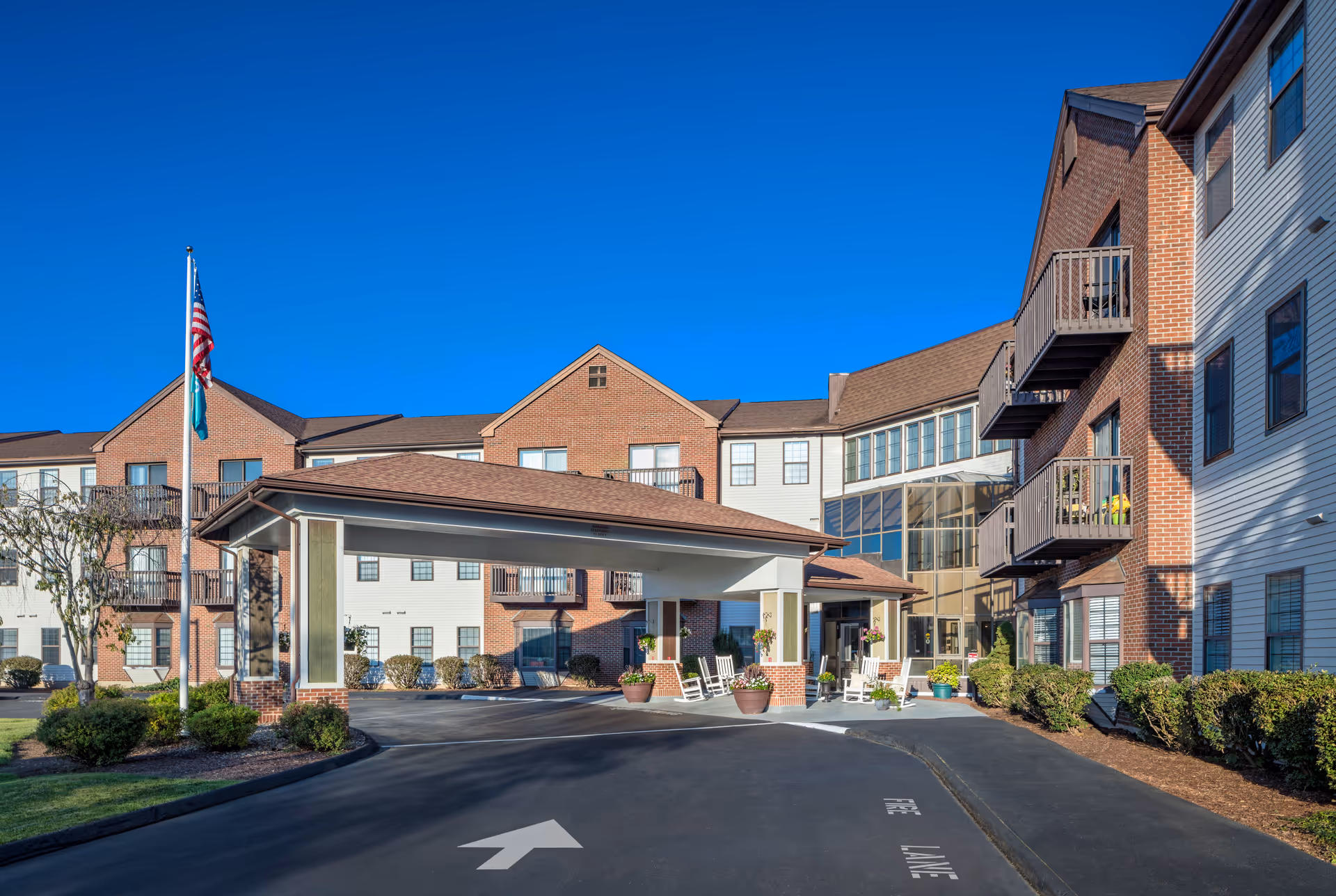 Exterior view of Cedar Woods senior living facility showing a three-story building with brick and siding facade, balconies, a covered entrance with rocking chairs, potted plants, and an American flag on a flagpole. The driveway has a white arrow and the words 'FIRE LANE' painted on the asphalt.