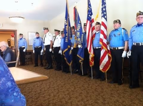 A group of veterans in uniform standing in a line indoors, holding various flags including the American flag, in a room with carpeted floor and white walls.