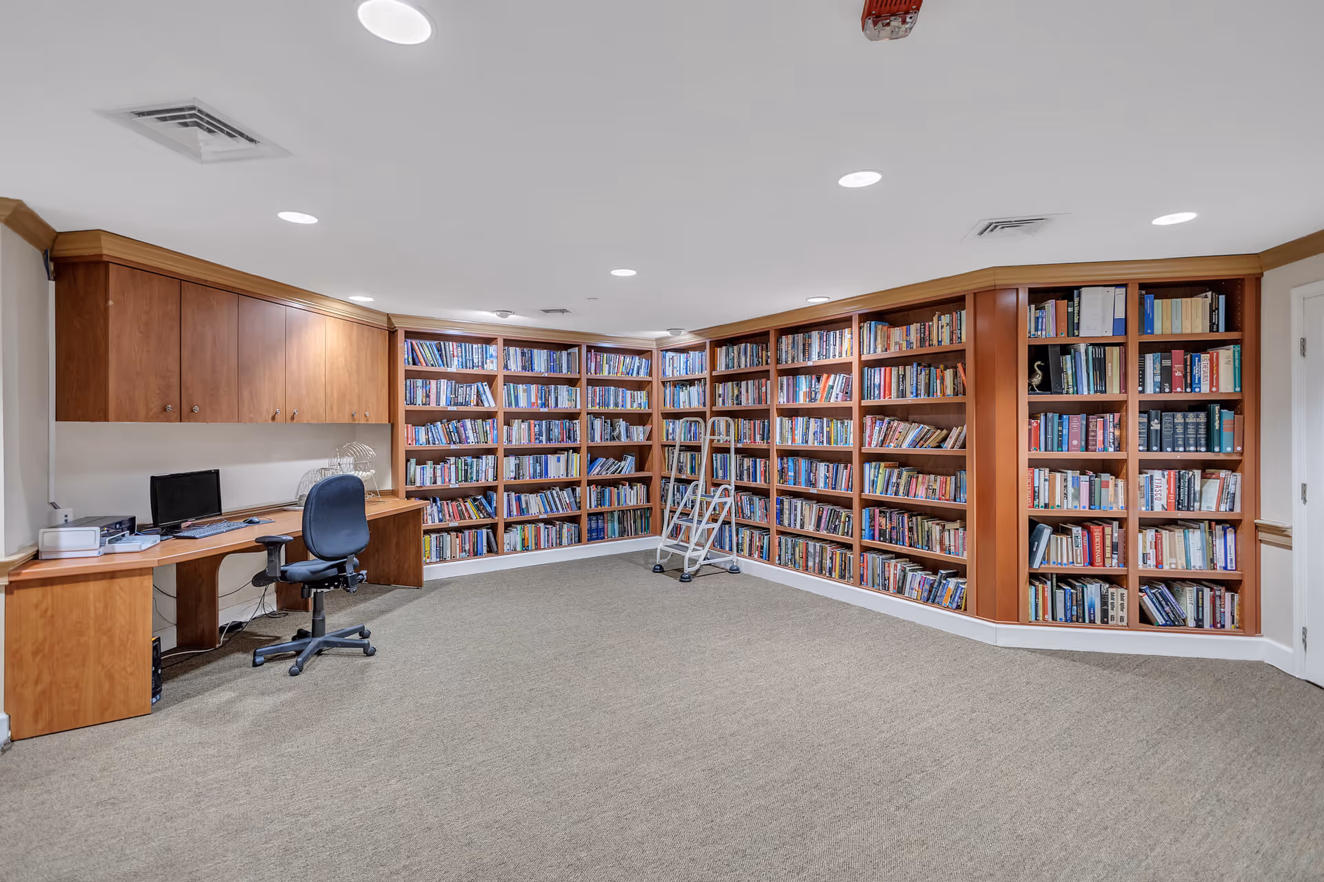 A spacious library room with carpeted floor and wooden bookshelves lining two walls, filled with numerous books. There is a small rolling ladder in front of the shelves for reaching higher books. On the left side, there is a built-in wooden desk with a computer, office chair, and overhead cabinets. The ceiling has recessed lighting and air vents.