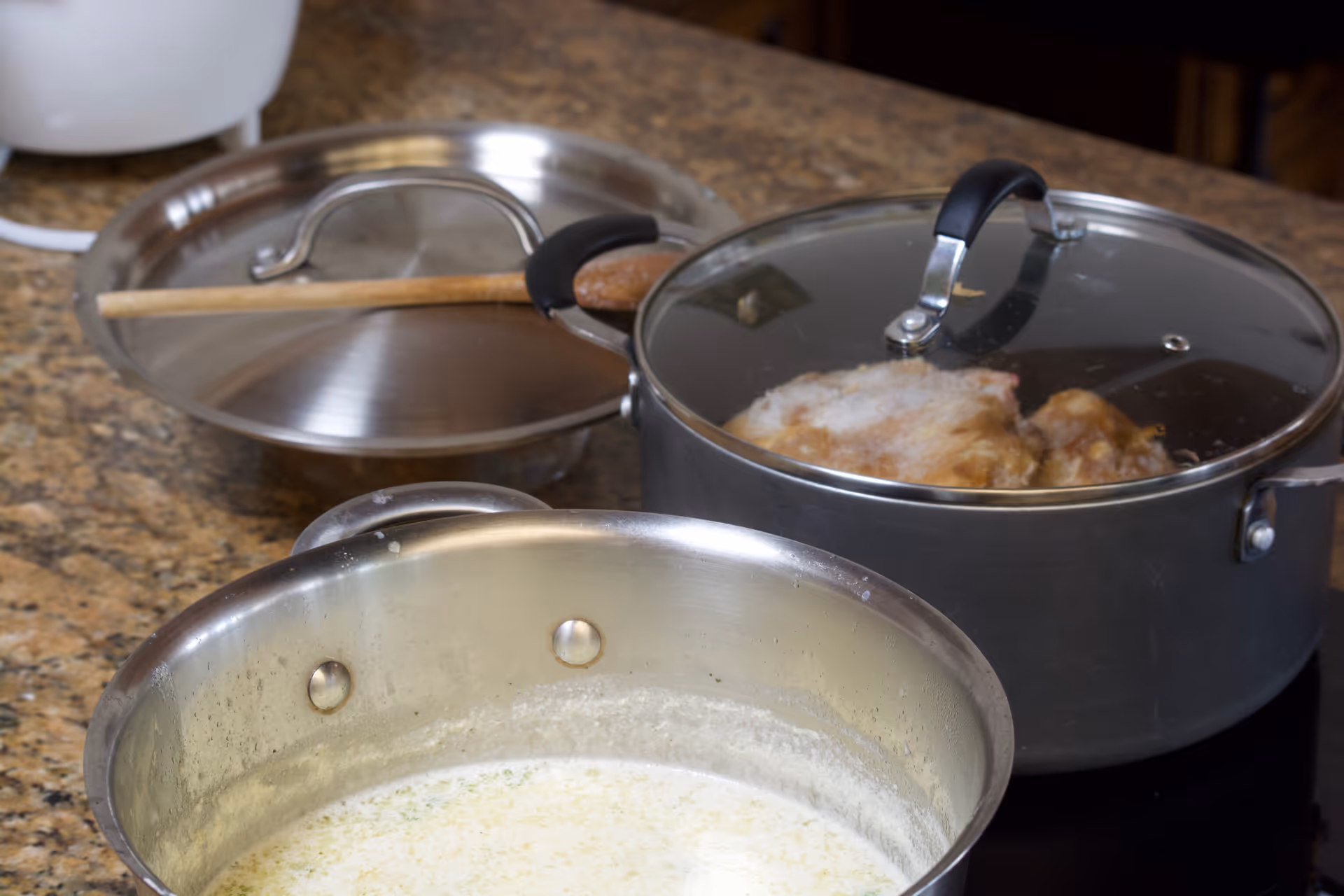 Close-up of two cooking pots on a kitchen countertop, one with a glass lid showing food inside and the other with a creamy sauce or soup. A wooden spoon rests on a metal lid in the background.