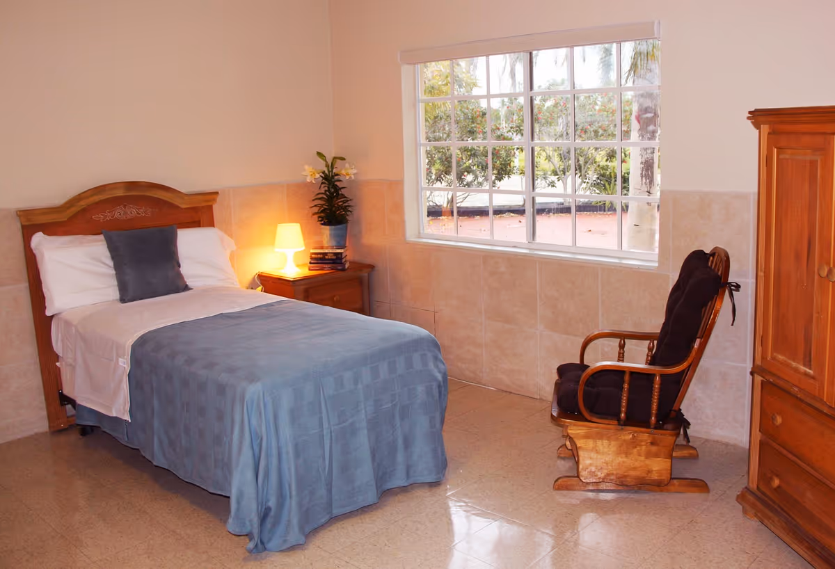 A simple, clean bedroom in an assisted living facility featuring a single bed with white sheets and a blue blanket, a wooden headboard, a nightstand with a lamp and a potted plant, a wooden rocking chair with cushions, a large window showing outdoor greenery, and a wooden wardrobe.