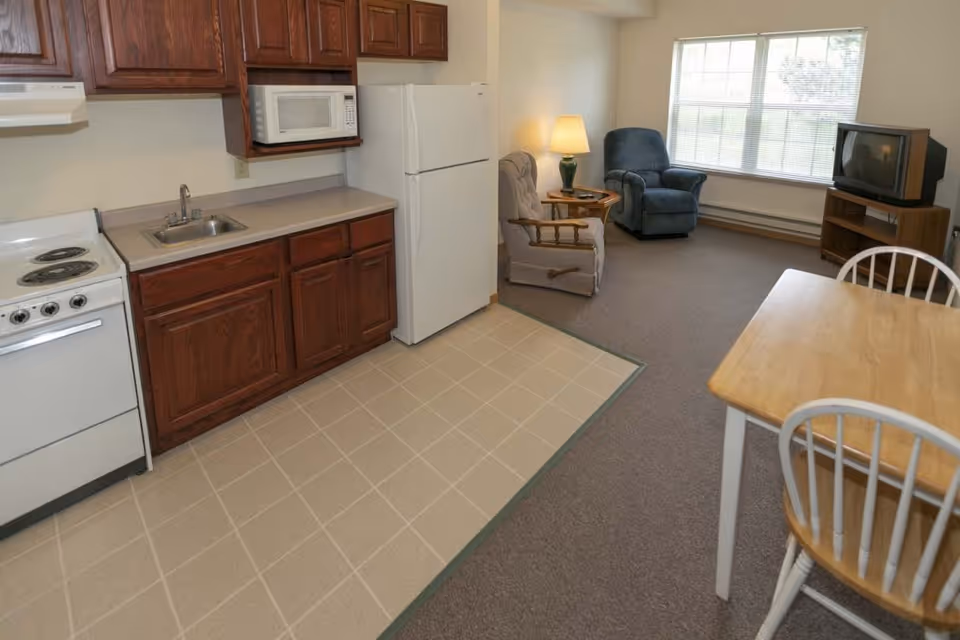 A small living space featuring a compact kitchen area with wooden cabinets, a white stove, microwave, and refrigerator. Adjacent to the kitchen is a dining table with two chairs. The living area includes two armchairs, a side table with a lamp, a large window, and an older-style television on a wooden stand.