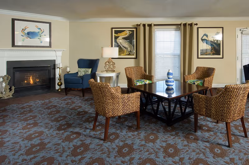Cozy common living room with wicker chairs arranged around a wooden coffee table, a fireplace, patterned carpet, and framed bird artwork.