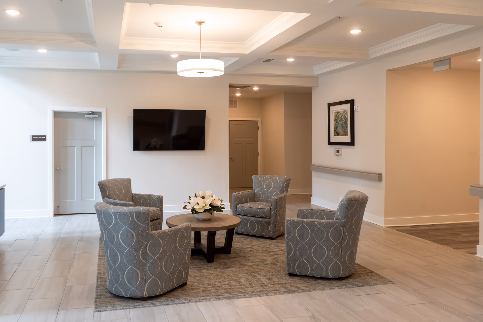 A modern lounge area with four gray patterned armchairs arranged around a round wooden coffee table with a white flower centerpiece. The room has light-colored tiled flooring, a large flat-screen TV mounted on the wall, recessed ceiling lights, and a framed artwork on the wall. There is a door labeled 'Office Manager' on the left side.