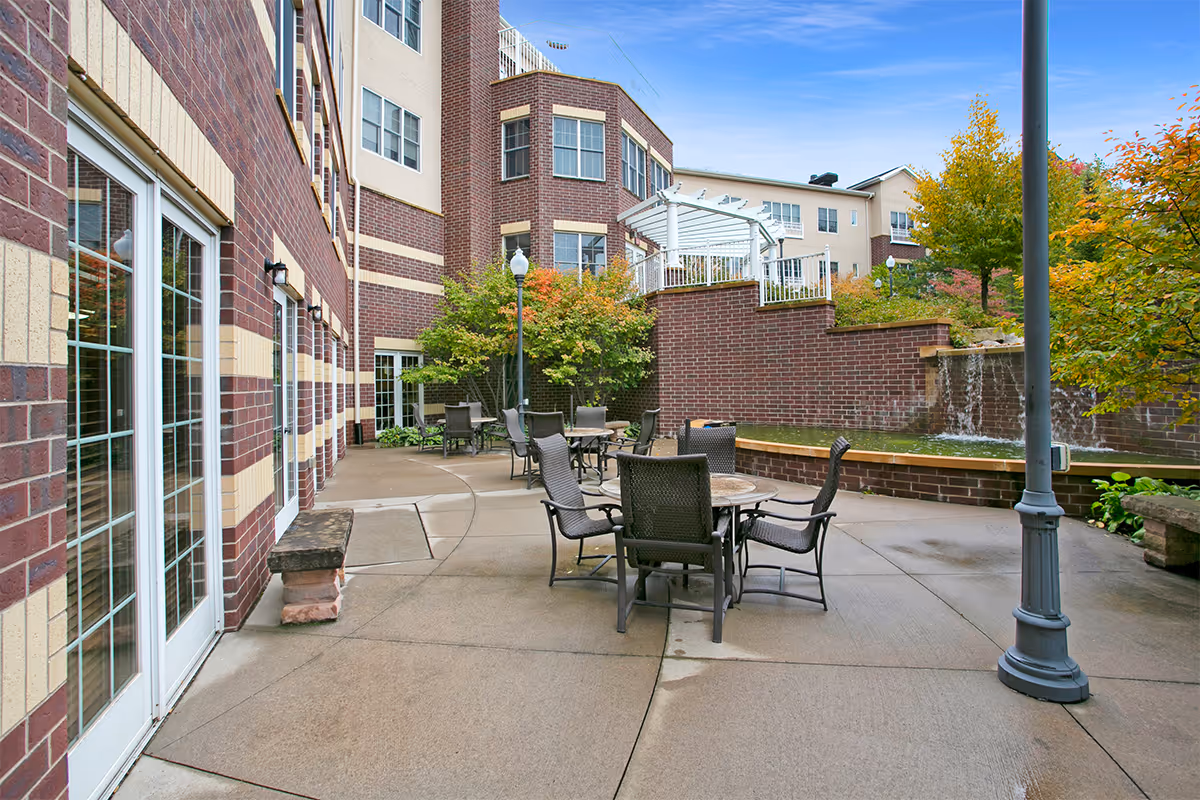 Outdoor patio area at Boutwells Landing featuring several round tables with chairs, a brick building with large windows, a lamppost, trees with autumn foliage, and a small waterfall feature on a brick wall.