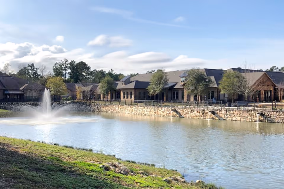 View of a senior living facility building named Watercrest at Kingwood, situated behind a pond with a water fountain in the center. The building is surrounded by trees and a stone retaining wall along the water's edge under a partly cloudy sky.