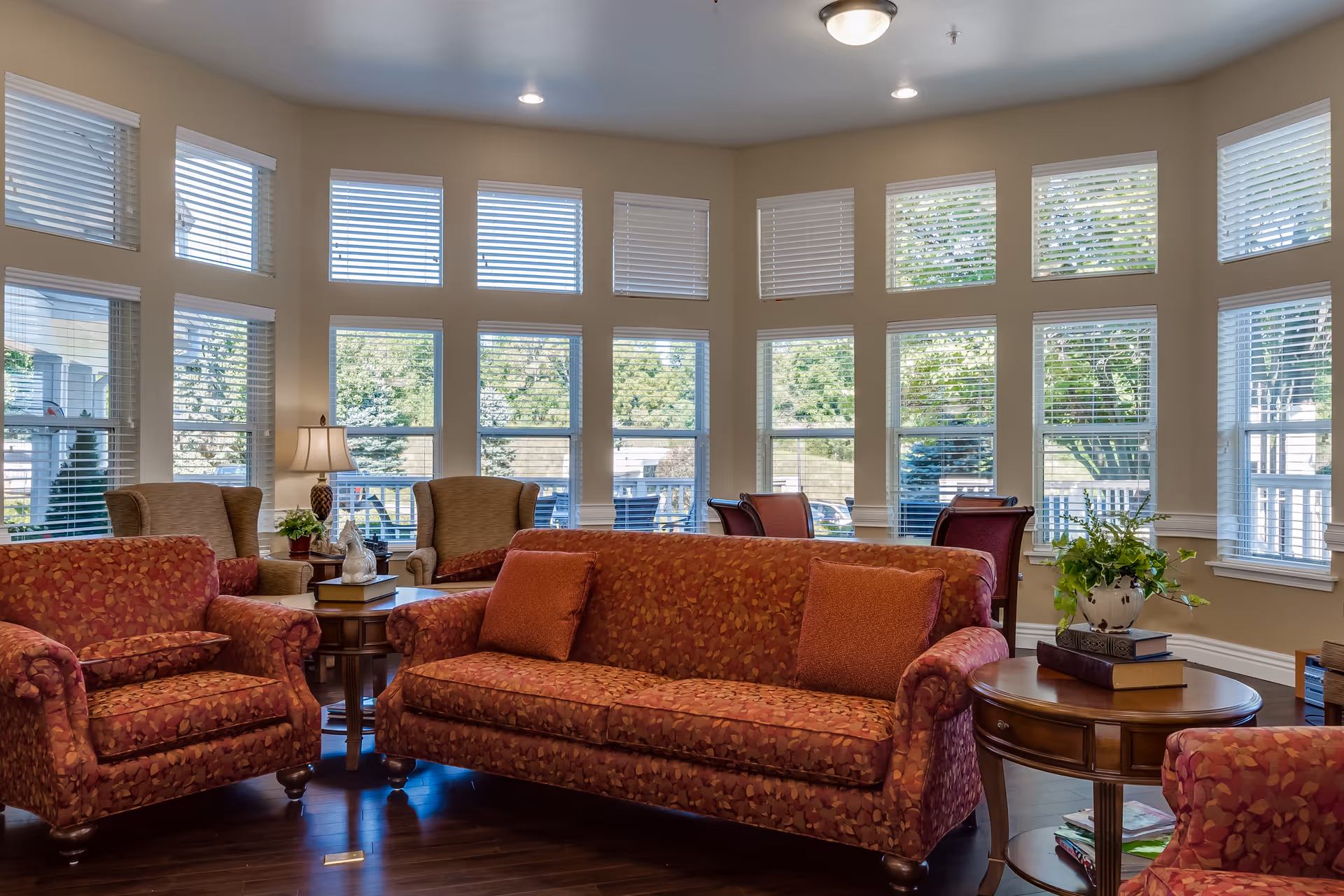 A bright and spacious living room with large windows letting in natural light. The room features patterned red sofas and armchairs, wooden side tables with books and a potted plant, and beige walls. Outside the windows, greenery and trees are visible.