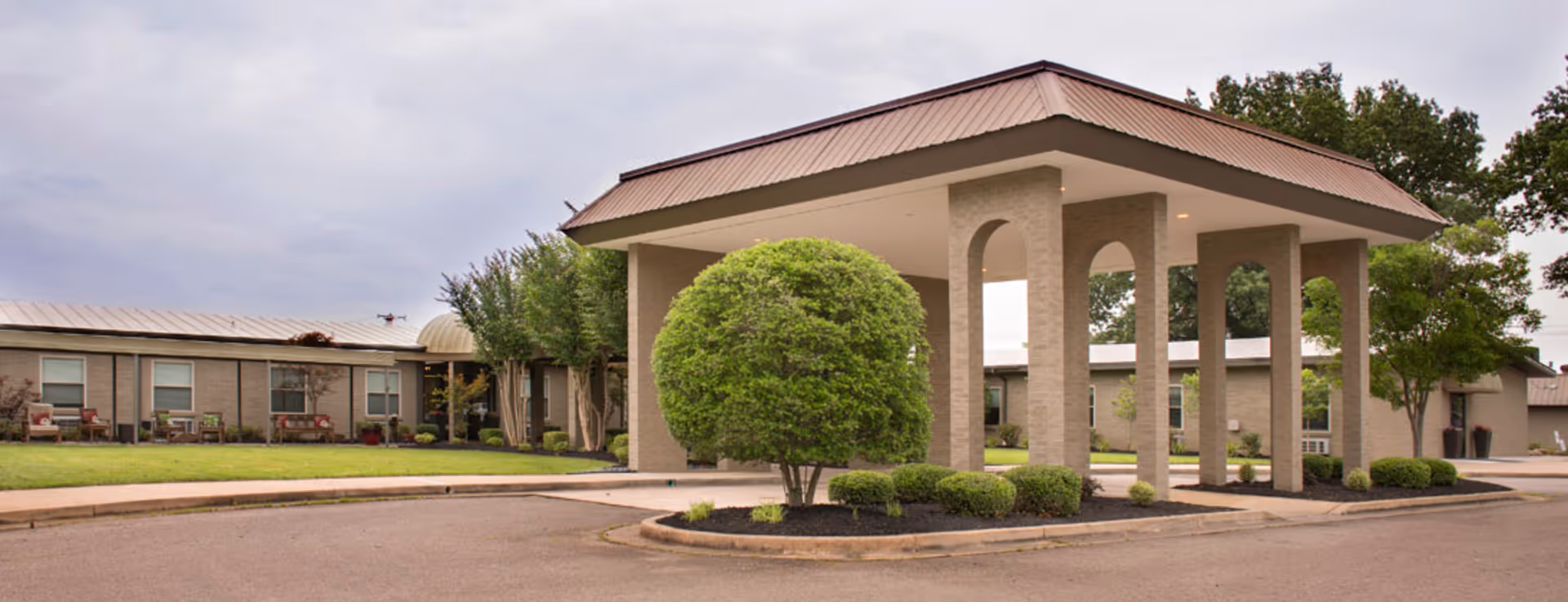 Exterior view of Stella Manor senior living facility showing a covered entrance with brick pillars, manicured bushes, and a lawn with benches in front of a single-story building under a cloudy sky.