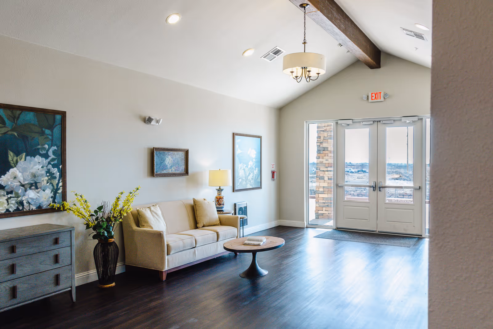 A bright assisted-living lobby with a beige sofa, coffee table, artwork, decorative vase, and double exit doors.